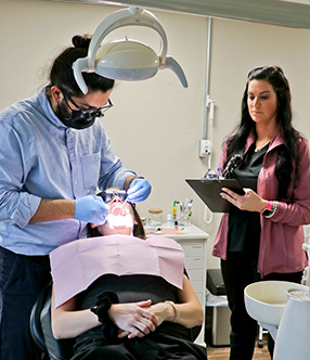 The image depicts a dental office setting where a dentist is performing oral care on a patient with the assistance of a dental hygienist, who is using a dental drill.