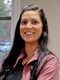 A smiling woman with long dark hair wearing a pink top stands against a window with a blurred background.