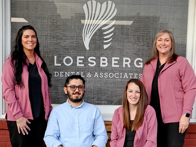 The image shows four individuals standing together outdoors, posing for a photograph with a sign that reads  LOESEBERG DENTAL ASSOCIATES  behind them.